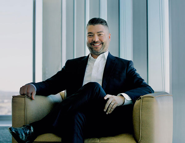 Kevin smiling wearing a suit and sitting in a chair with skyline behind him